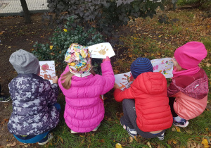Dzieci kucają pod drzewem, w rękach trzymają swoje prace plastyczne.